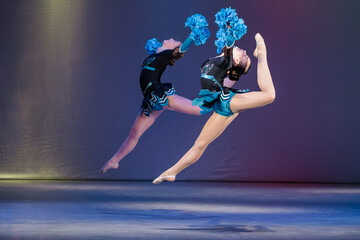 Two young cheerleaders perform a synchronized jump with high leg extensions, holding blue pom-poms mid-air. Their dynamic pose, strength, and flexibility are highlighted against a colorful background. © ig_royal