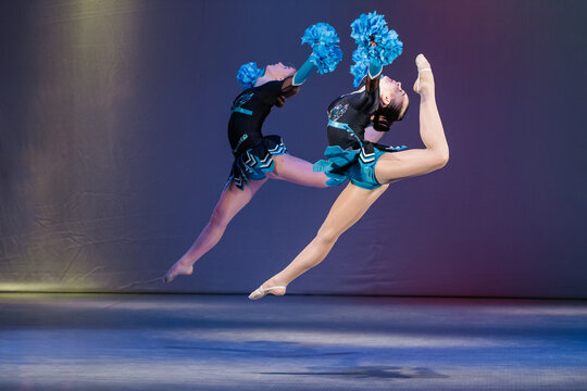Two young cheerleaders perform a synchronized jump with high leg extensions, holding blue pom-poms mid-air. Their dynamic pose, strength, and flexibility are highlighted against a colorful background.