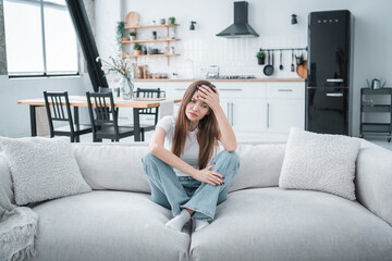 Fototapeta premium Young tired unhealthy Caucasian woman sitting on couch in living room at home holding head with hand suffering from strong sudden headache or migraine throbbing pain.