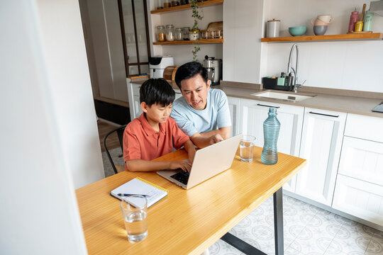 Chinese father guiding son with laptop in modern kitchen