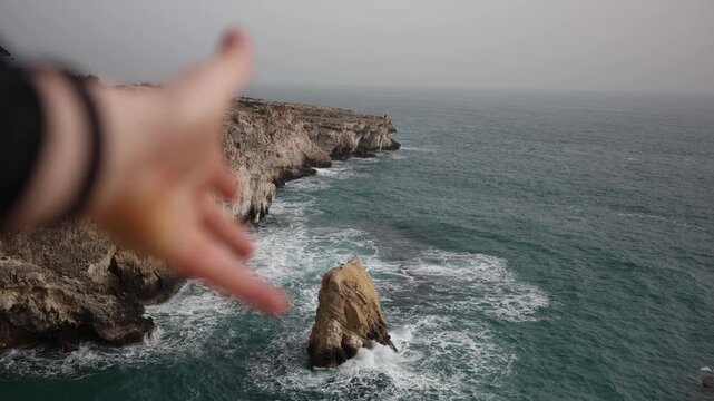 Outstretched hand reaching toward rugged sea cliffs and crashing waves cinematic coastal freedom wind mist wild ocean travel feeling