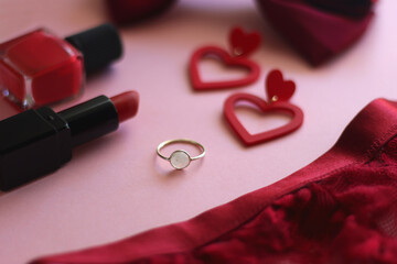 Red lace bralette, hairbow, heart shaped earrings, golden ring, red lipstick and nail polish on pink background. Coquettish accessories for Valentine's day. Selective focus.