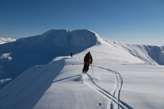 Off-piste freeride skiers traverse a snow-covered ridge under a clear blue sky in fresh powder on a high alpine peak