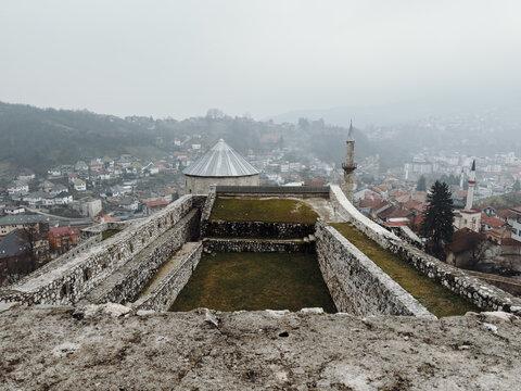 View from medieval fortress walls overlooking Travnik city in dense fog, showing stone defenses, rooftops, and layered hills under an overcast sky.
