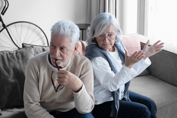 Fototapeta premium Elderly couple sitting back to back on couch having a dispute, expressing relationship conflict, unresolved problem or question, emotional distance, or communication problems in a modern living room.