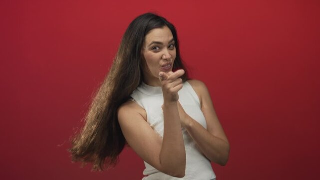 Woman wearing white sleeveless top smiling and pointing index finger toward camera in studio with red wall; confidence.