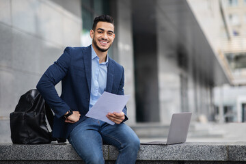 Handsome arab entrepreneur sitting by office building with laptop, holding documents and smiling, copy space. Confident cheerful guy in suit getting ready before business meeting, reading speech