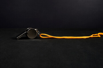 A silver metal whistle suspended by a yellow braided cord against black background, symbolizing...