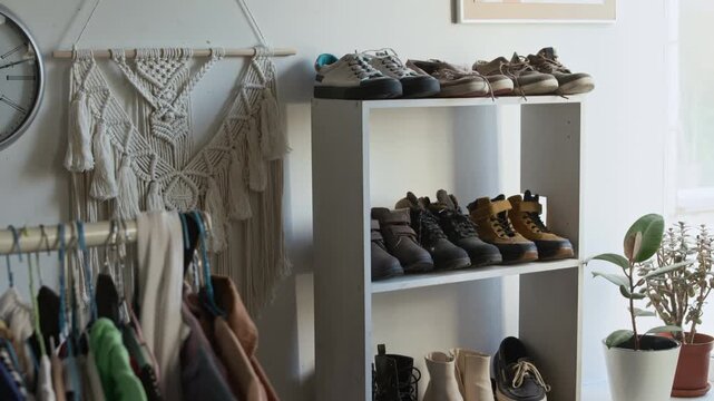 Medium panning shot of assorted preowned multicolored womenswear hanging on rack, boots and shoes displayed on shelves in charity shop, macrame piece decorating wall, conscious consumption vibe