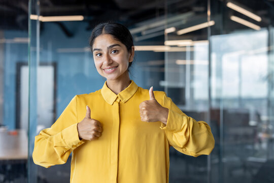 Portrait of a young Indian woman standing in the office and showing the camera a successful super sign with her fingers