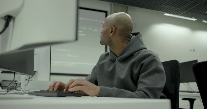 Concentrated Man coding in a modern tech scene, wearing Hoodie, Hacker, looking at computer code on monitors in office