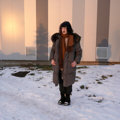A stylish young woman with dark hair and bold makeup stands outdoors in a snowy environment, wearing a long gray puffer coat with a fur-trimmed hood, a brown scarf, black pants, and black boots. © Dina Luk