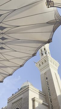 A stunning low-angle view of the iconic umbrellas and towering minarets at Al-Masjid an-Nabawi (The Prophet's Mosque) in Medina, showcasing beautiful Islamic architecture against a clear blue sky.