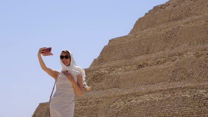 A woman taking a selfie beside an ancient Egyptian pyramid, enjoying a sunny vacation and travel moment in Egypt.
