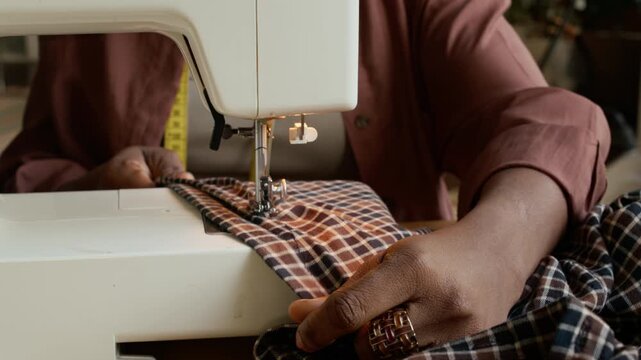 Close-up of hands of unrecognizable black female seamstress sewing garment from swath of checked fabric on machine, while working in atelier or second hand shop, recycling preowned clothing