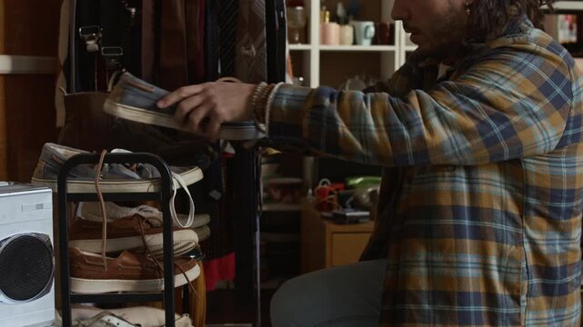 Medium side shot of young Caucasian man searching through preowned leather boots and canvas shoes displayed on rack at charity outlet, checking size and style, suggesting thrifty shopping