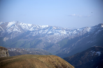 Obraz premium Snow-capped mountains under a clear blue sky, with a small village nestled in the valley. Rolling hills in the foreground, creating a serene and expansive landscape.