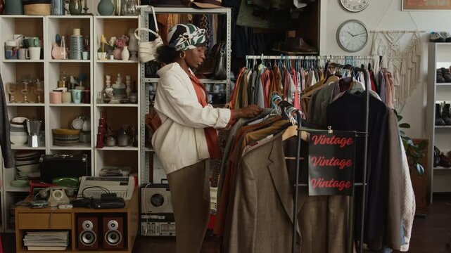 Medium shot of multiethnic young people shopping for bargains at second hand store, Black woman examining blouse on hanger in womenswear, Caucasian man taking smartphone photos of homeware on shelf