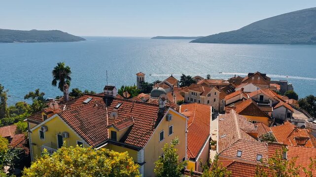 Montenegro, picturesque scenery of seaside promenade on a sunny day, moored boats, brick-colored tiled roofs, a mountain range in the background