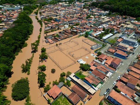 Vista a&eacute;rea por drone de uma enchente urbana, mostrando ruas alagadas, casas parcialmente submersas e o impacto das chuvas intensas. Conceito de desastre natural, mudan&ccedil;as clim&aacute;ticas, crise ambiental.