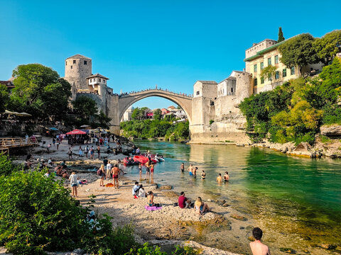 Old town of Mostar with historic stone houses, traditional bridge and turquoise river surrounded by green hills and mountains under clear summer sky.