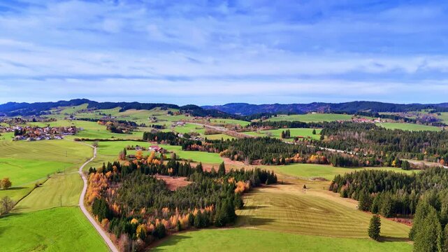 Green fields and forests in a rural landscape with blue sky and clouds. Fields and forests span the landscape with homes dotting the area under a cloud-filled sky.