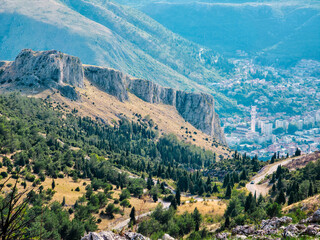 Rocky hills and forested slopes above a Bosnian town. Mountain landscape with limestone cliffs, winding road and dense greenery under clear summer sky.