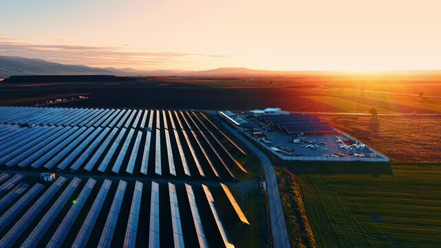 Solar panels gather sunlight during sunset near a field and building. Rows of solar panels capture sunlight as the sun sets behind hills and fields near a building.