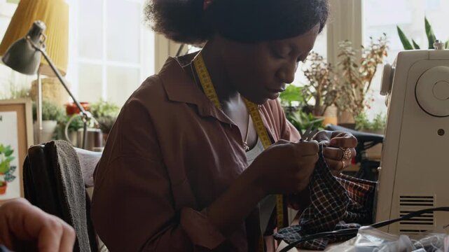 Medium shot of busy young African American seamstress removing stitches on preowned textile garment for upcycling, chatting to friend or colleague, sewing together in workshop of second hand outlet