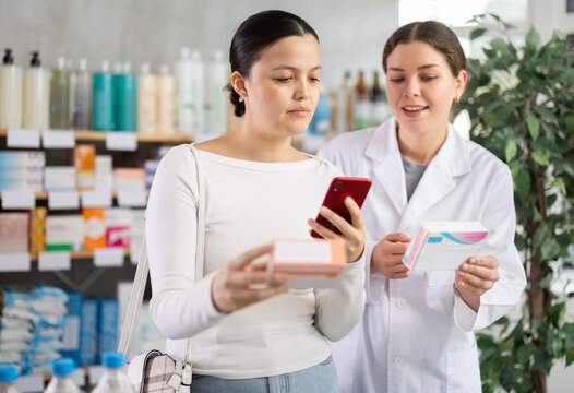 Thoughtful female customer using smartphone to scan medications QR code, checking details while friendly pharmacist giving professional advice at local pharmacy