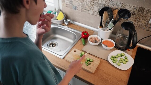 Focused teenager skillfully slicing fresh cucumber with knife on wooden cutting board, prepare healthy meal and tasting piece in modern kitchen with natural light