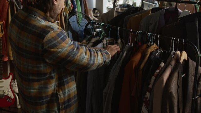 Medium shot of young Caucasian male hipster with dreadlocks browsing vintage jackets displayed on rack at charity shop, searching for bargain fashion clothes, conscious consumption vibe
