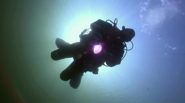 Lone diver, illuminated by bright flashlight, swims through clear, cool waters of White Sea. Sunlight streams through water's surface above, creating surreal and breathtaking aquatic ambiance.