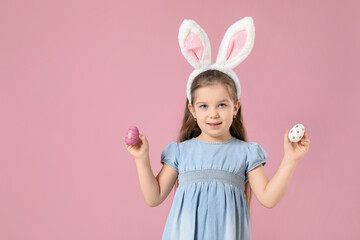 Easter celebration. Smiling little girl in headband with bunny ears holding painted eggs on pink...