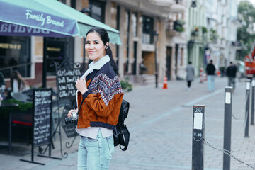 Obraz premium Street scene with a woman wearing a patterned jacket and light jeans, carrying a backpack, smiling softly as she strolls past cafe terraces on a sunny urban day.
