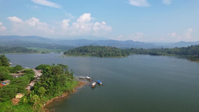 Aerial drone footage of Jatigede reservoir, with flat waters, some islands with forests, fishing boats, houses and some stalls, in sunny weather, in Sumedang regency, Java island, Indonesia 