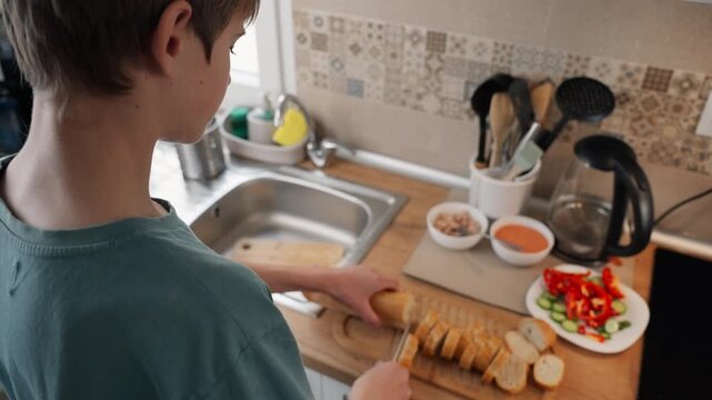 Young teen boy slicing fresh baguette with knife on wooden cutting board in kitchen, prepare healthy meal with fresh vegetables and ingredients