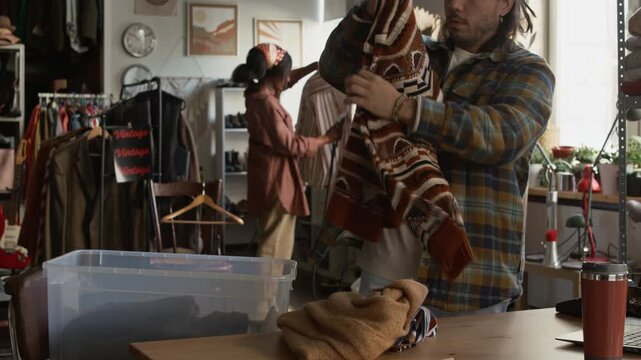 Medium shot of young Caucasian male charity shop worker standing by counter, checking out and sorting donated sweaters, making notes on laptop and preparing garments for sale
