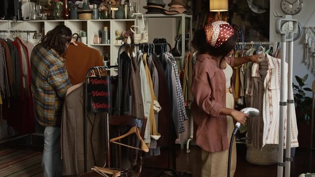 Medium shot of busy young African American female second hand shop worker steam ironing creases on preowned shirt, preparing for sale, while Caucasian male colleague checking clothes on rack