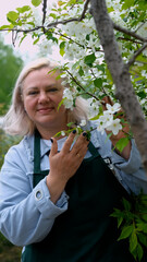 A portrait of a happy woman gardener, standing next to a blossoming apple tree, smiling, enjoying the pleasant scent of the flowers, and feeling happy.