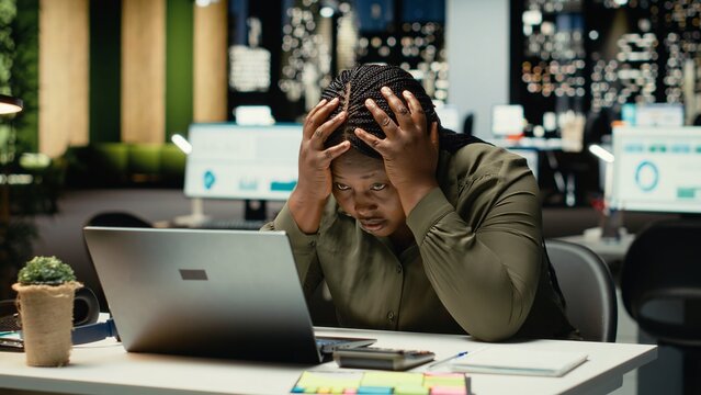 Black restless weary woman experiencing burnout and fatigue late at night, being overwhelmed and overworked at desk. Anxious fatigued person struggling with mental haze, discomfort. Camera B.