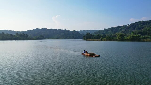 Aerial drone footage at sunset, of a small wooden fishing skiff, loaded with wood and 2 people, in flat waters of the Jatigede reservoir, with forests and hills behind, in Sumedang regency, Indonesia