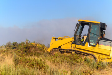 A yellow bulldozer clears vegetation to create a firebreak during a forest fire © VicVaz