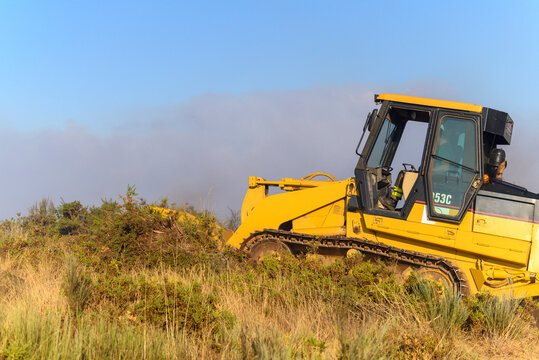 A yellow bulldozer clears vegetation to create a firebreak during a forest fire