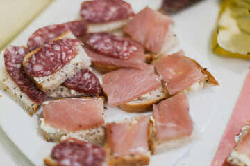 A close-up shot displays an assortment of small appetizers, featuring slices of bread topped with various deli meats, neatly arranged on a white serving platter.