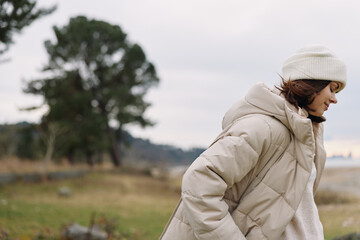 Woman in a beige puffer coat walks outdoors on a breezy day with a knit hat, in a natural setting of trees and open field, conveying seasonal fashion and calm © SHOTPRIME STUDIO