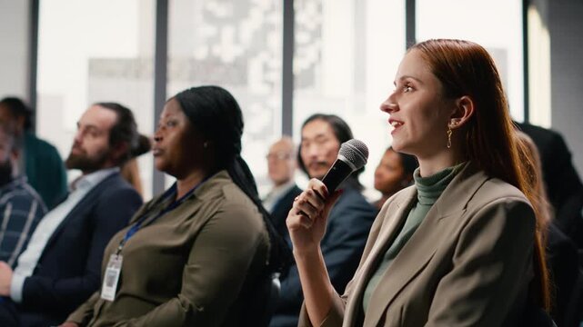Female participant raising hand and engaging in debate on microphone, attending corporate summit in conference hall with chairs and active audience. Diverse group at professional event. Camera B.