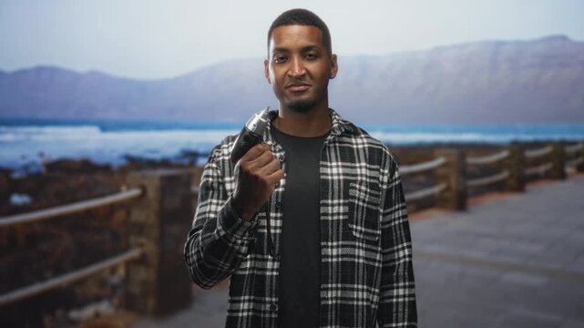 Man holding electric screwdriver in right hand, pointing its tip slightly forward while standing on a street boardwalk by the sea with wooden railings; confidence readiness.