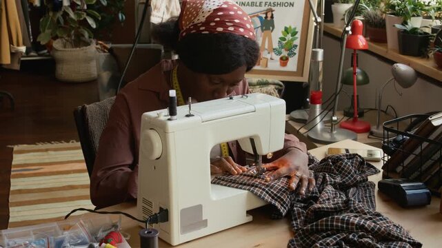 Medium pan shot of two diverse second hand shop employees upcycling preowned textiles while working together, sewing garment from checked fabric and mending jacket with elbow patches by hand