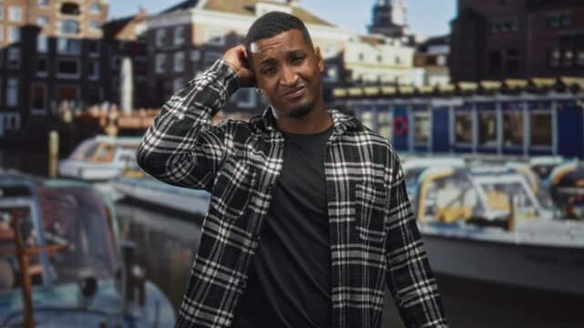 Young man scratches head, hand to temple gesture before canal boats backdrop in studio; uncertainty contemplation.